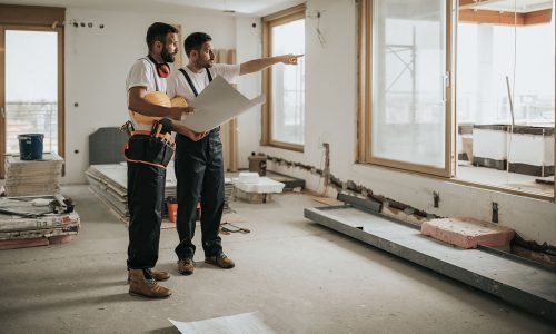 Full length of construction workers analyzing blueprints in the apartment while one of them is aiming at distance.